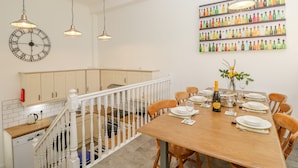A dining area with a wooden table set for six, white chairs, and a chandelier. A large clock and a colorful bottle shelf are visible on the walls.