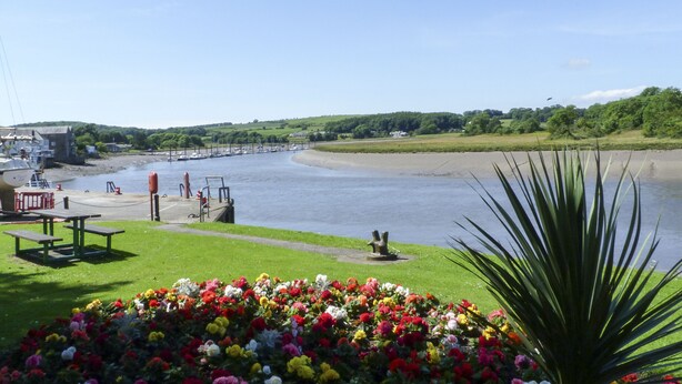 Une vue sur la rivière, avec un espace verdoyant, des bancs et un parterre de fleurs.