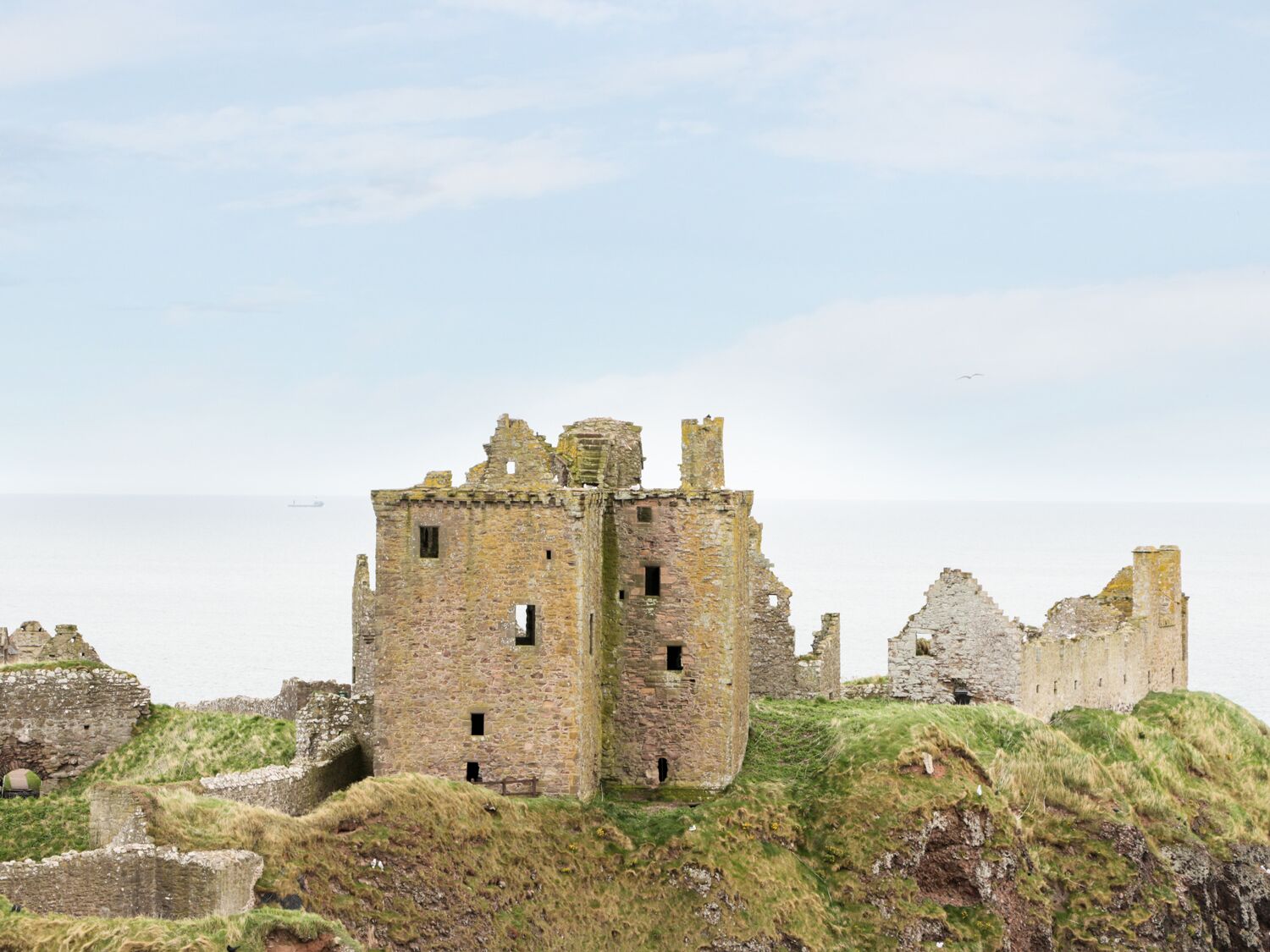 Rovine di un castello in pietra di epoca storica, con numerose torri e mura, arroccato su una collina erbosa con vista sul mare.
