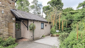 A stone building with a tiled roof, a small porch, and a garden area with a hanging flower basket. - Doe Foot Cottage (Carnforth)