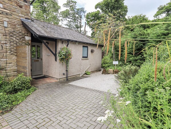 A stone building with a tiled roof, a small porch, and a garden area with a hanging flower basket. - Doe Foot Cottage (Carnforth)