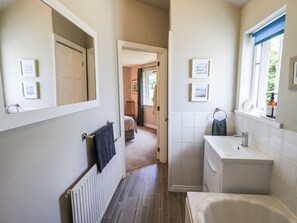A narrow bathroom with a bathtub, a sink, and a mirror. There are framed pictures on the walls and a towel hanging on the radiator. - Doe Foot Cottage (Carnforth)
