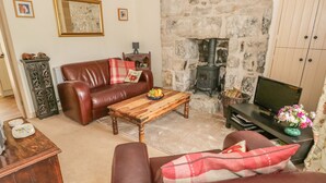 A cozy living room with a stone fireplace, leather sofa, wooden coffee table, and a television. - Herdwick Cottage (Grange-over-Sands)