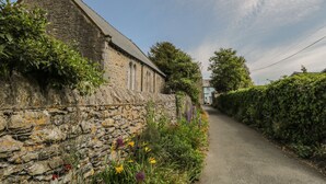 A stone building with a sloped roof, a narrow road beside it, and a hedge along the road. - Herdwick Cottage (Grange-over-Sands)