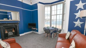 A room with a blue wall, a fireplace, a TV, a dining table with chairs, and a leather sofa. - Apartment 3 (Tenby)