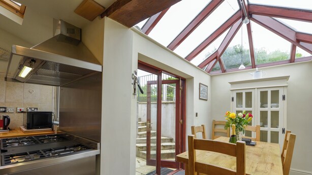 A kitchen with a gas stove, a microwave, and a stainless steel range hood. There is a dining area with a wooden table and chairs, and a glass door leading outside. - 11 The Village (Yelverton)