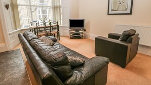 A living room with a leather sofa, a small table with chairs, a flat-screen TV, and a framed artwork on the wall. - Flat 4 (Whitby)