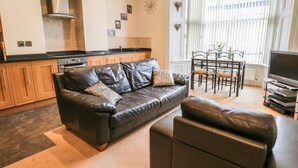 A modern living room with a black leather sofa, a dining table with chairs, a flat-screen TV, and a kitchen area with wooden cabinets and a stove. - Flat 4 (Whitby)