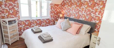 Un dormitorio con cama, almohadas, una mesita de noche, una lámpara, una ventana y una pared con estampado floral.