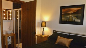 A bedroom with a bed, a bedside table with a lamp, a mirror, and a bathroom with a sink. - Daisy Cottage (Penrith)