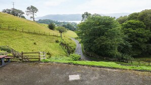A rural road curving through a green landscape with a fence on one side and a wooden gate at the entrance. - Low Skelgill (Windermere)