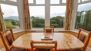 A round wooden table with six chairs, a book, and a view of a green landscape through large windows. - Low Skelgill (Windermere)