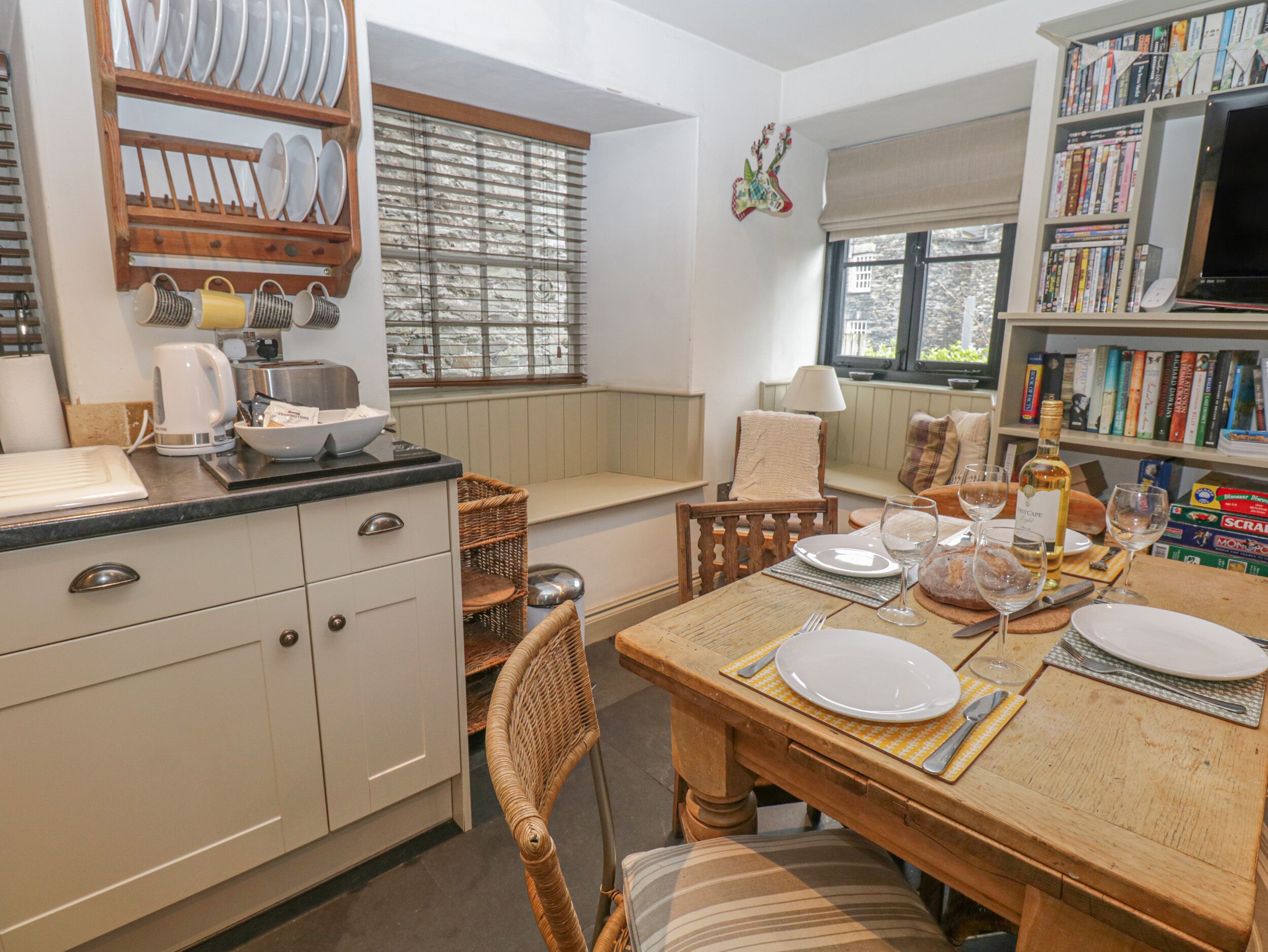 Una cocina acogedora con una mesa de comedor dispuesta para dos, una estantería llena de libros y una ventana con persianas.