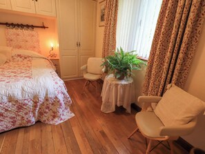 Ein Schlafzimmer mit floralem Bettbezug, einem Sessel, einem kleinen Tisch mit Pflanze und einem Fenster mit Vorhängen.