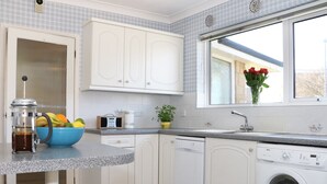 Une cuisine moderne avec des meubles blancs, une machine à café, un saladier rempli de fruits, un lave-linge et une fenêtre donnant sur un beau paysage.