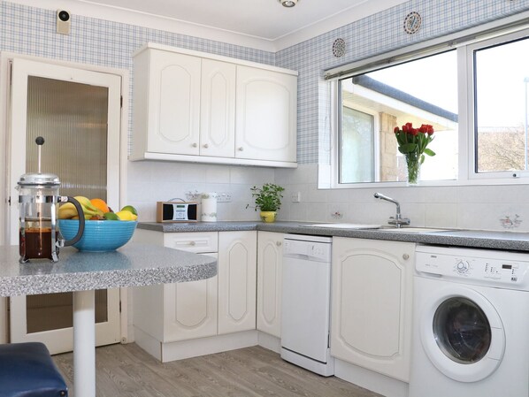 A modern kitchen with white cabinets, a coffee maker, a bowl of fruit, a washing machine, and a window with a view.