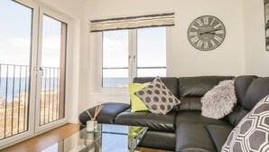 Uma sala de estar com sofá de couro preto, mesa de centro de vidro e uma grande janela com vista para o mar. É visível um relógio de parede e há uma pequena mesa com uma vela e difusores de vagem.
