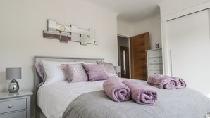 Cama bem arrumada com cobertor cinza e toalhas enroladas, luminária de cabeceira e um objeto decorativo na parede acima da cama.
