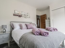 A neatly made bed with a grey blanket and rolled towels, a bedside lamp, and a decorative wall piece above the bed.