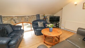 A modern living room with a stone wall, a black leather sofa, a blue armchair, and a wooden coffee table with a vase of flowers. - The Old Bakery Barn (Bakewell)