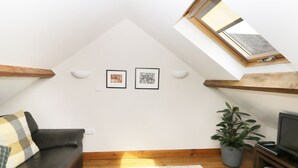 Ein Loft-Zimmer mit schrägverdeckter Decke, Holzbalken, einer schwarzen Leder-Couch, einem weißen Teppich, einer Topfpflanze und einem Fernseher auf einem Holzgestell.