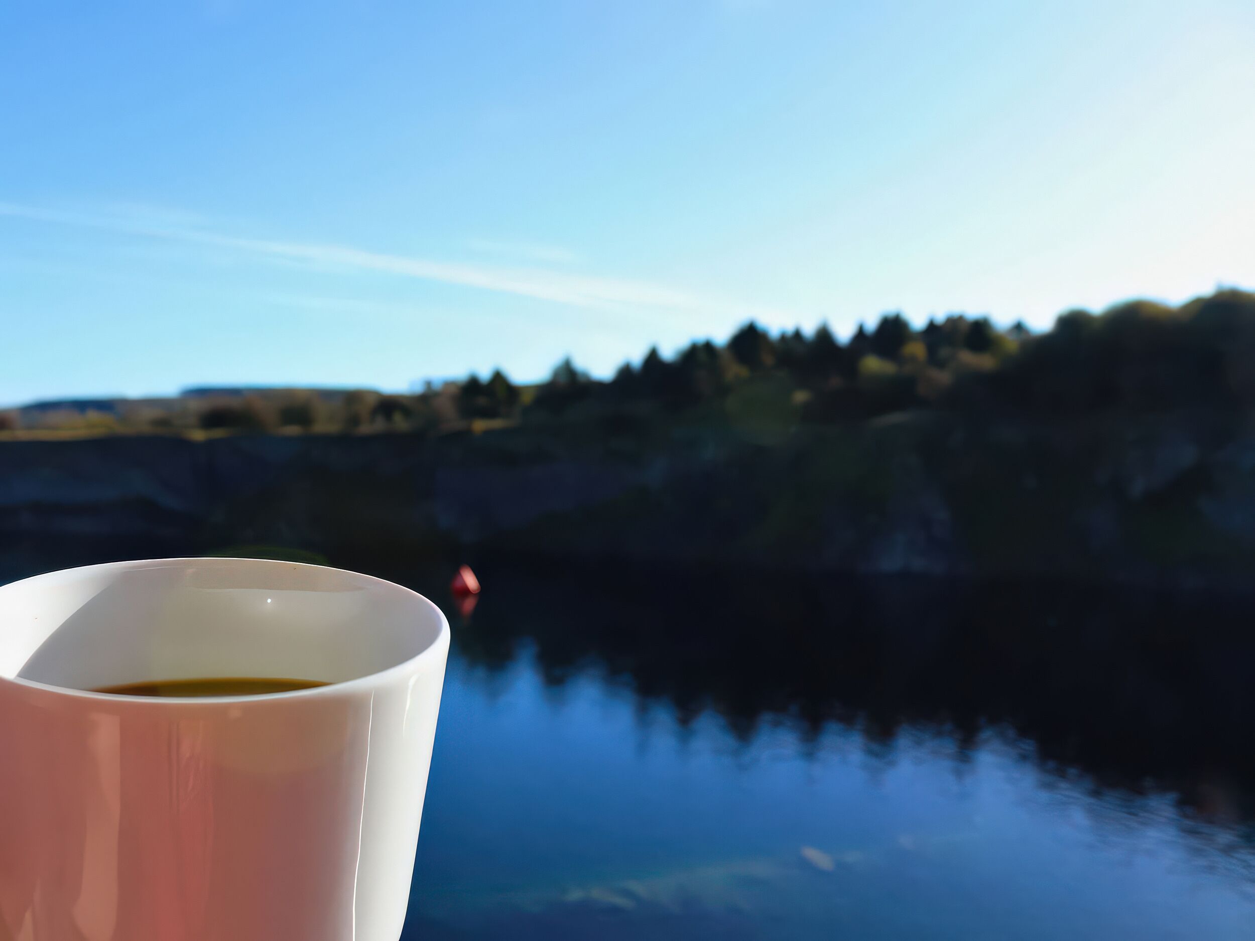 Une tasse blanche au premier plan, avec un arrière-plan flou représentant une étendue d’eau calme et une colline boisée.