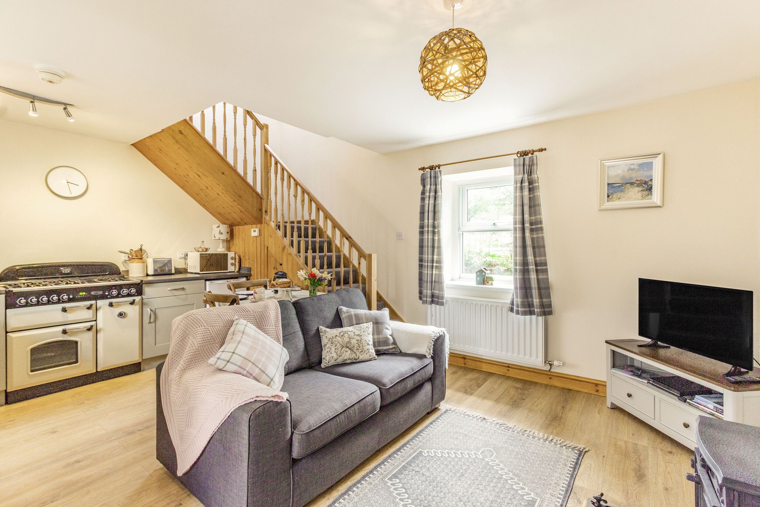 A cozy living room with a grey sofa, wooden staircase, kitchen area, and a TV.