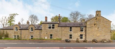 A stone building with multiple chimneys, a paved road in front, and a clear sky.