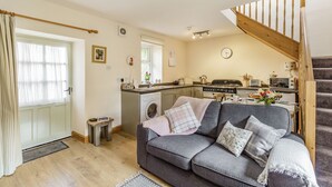 A cozy living room with a grey sofa, a wooden staircase, a kitchen area, and a washing machine. - Briar Cottage (Alnwick)