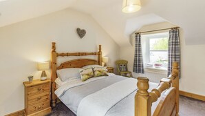 A bedroom with a wooden bed, a nightstand with a lamp, a chair, and a window with curtains. - Briar Cottage (Alnwick)