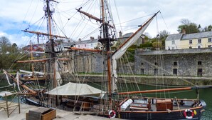 Un navire à voile historique est amarré dans un port, avec un bâtiment en pierre en arrière-plan.