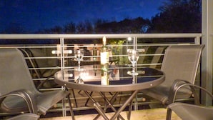 Une table ronde en verre, accompagnée de deux chaises, d’une bouteille et de deux verres à vin, est placée sur un balcon extérieur d’où l’on peut admirer des arbres et un ciel sombre.