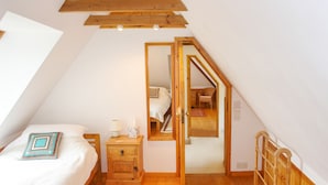 Un dormitorio con techo inclinado, vigas de madera, una cama, una mesita de noche y un espejo, con vista a otra habitación.