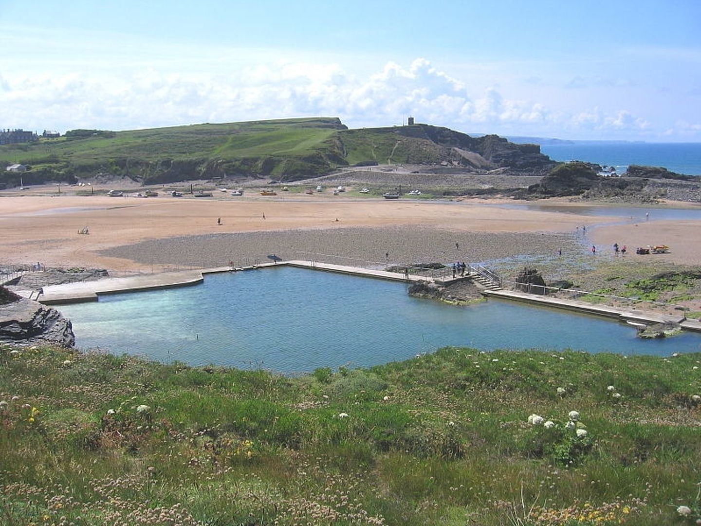 A coastal landscape with a large body of water, a sandy beach, and a grassy hill with a structure on top.
