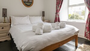 A neatly made bed with white linens and towels, a wooden headboard, a nightstand with a lamp, and a window with floral curtains. - Mollys Cottage (Penzance)