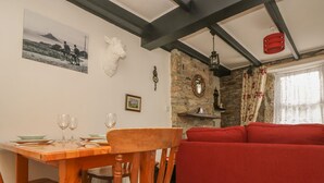 A dining area with a wooden table and chairs, a red sofa, and a stone wall with a mirror and shelf. - Mollys Cottage (Penzance)