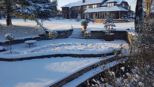 Un paisaje nevado con un puente de piedra, una terraza de madera y una casa grande con techo cubierto de nieve.