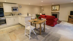 Uma cozinha moderna com armários brancos, micro-ondas e forno embutido. Há uma sala de jantar com mesa de madeira e cadeiras brancas. Um sofá vermelho está na sala de estar, e uma lareira é visível ao fundo.