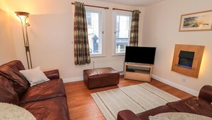 A living room with a brown leather sofa, a television on a wooden stand, a fireplace, and a window with curtains. - Seahorse (Whitby)
