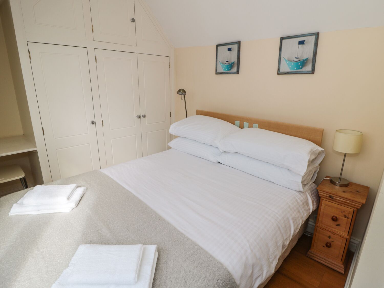 A neatly made bed with white linens, a wooden nightstand with a lamp, and two framed pictures on the wall.