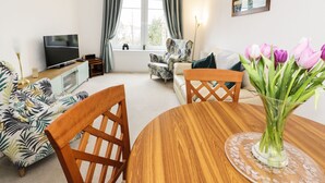 Una sala de estar con televisor, sofá, sillas, una mesa de comedor y un florero con flores.