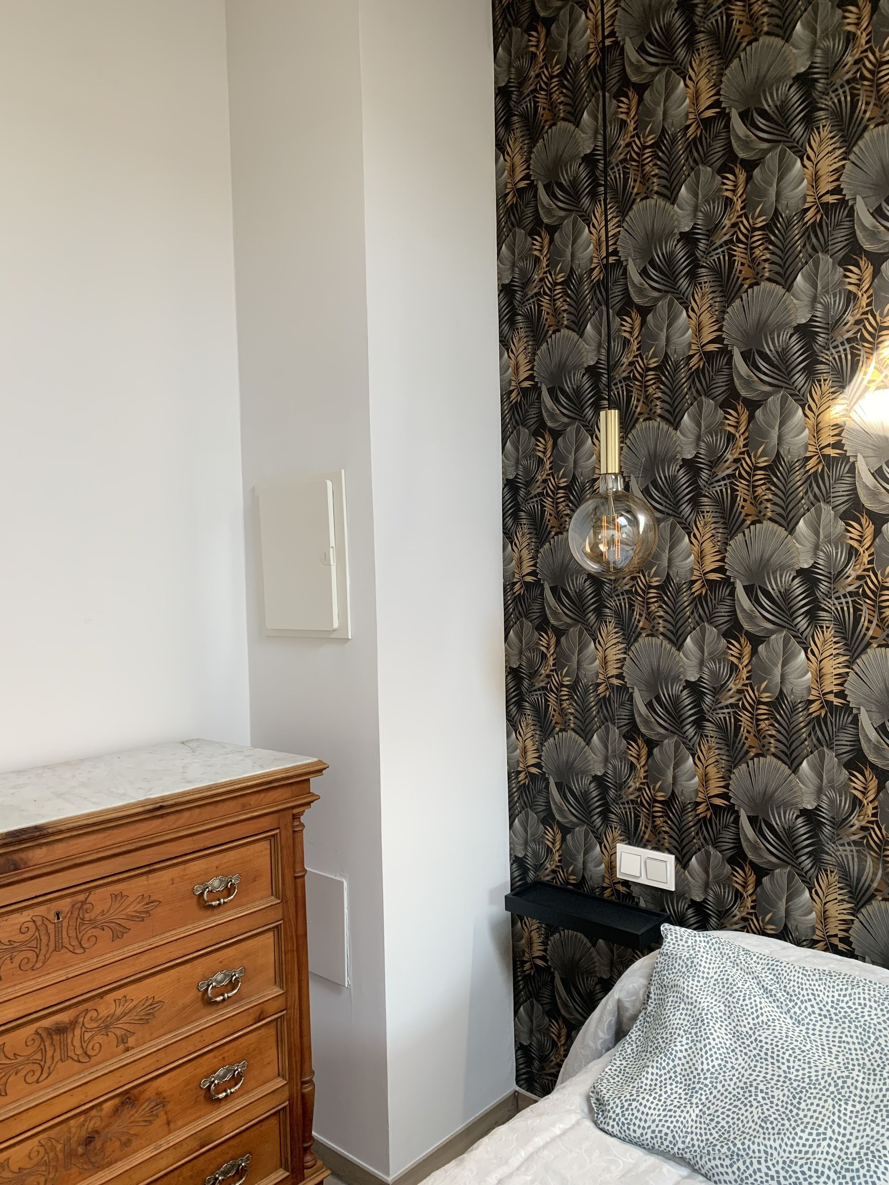 Un dormitorio con un aparador de madera, una cama con un cojín estampado y un papel tapiz con motivos de hojas.