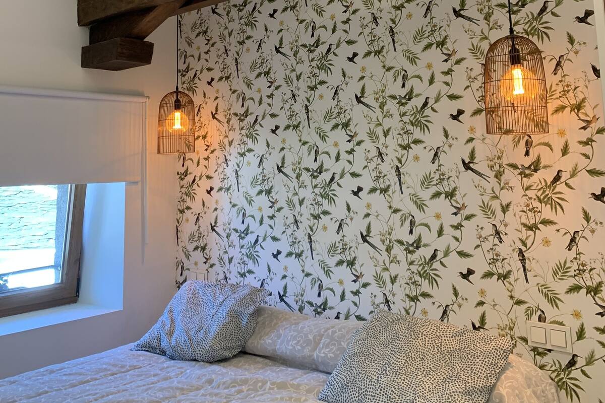 Ein Schlafzimmer mit einem Bett, strukturiertem Wandpapier, Holzbalken und zwei Hängeleuchten.