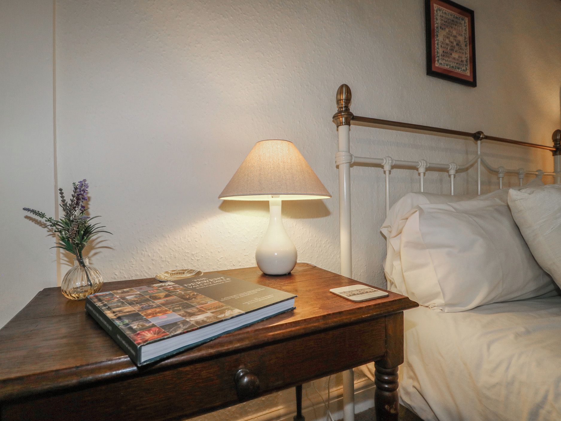 A neatly made bed with white linens, a bedside lamp, a book, and a small vase with flowers on a wooden nightstand.