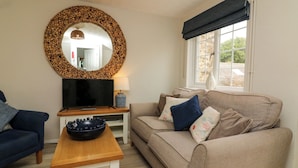 A living room with a beige sofa, a blue armchair, a wooden coffee table, a TV on a stand, and a large mirror with a wood frame. - Martin Cottage (Ilfracombe)