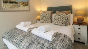 A neatly made bed with a plaid blanket, two pillows, and rolled towels. There is a painting of a beach scene on the wall, a bedside lamp, and a small wicker basket on the nightstand. - Martin Cottage (Ilfracombe)