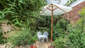 A garden patio with two white metal chairs under a white umbrella, surrounded by lush greenery and potted plants.