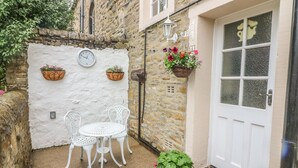 Un piccolo patio con tavolo e sedie bianche, un muro bianco con un orologio e cesti di fiori pensili.