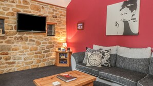 A cozy living room with a stone wall, a flat-screen TV, a wooden coffee table with books, a grey sofa with patterned cushions, and a red accent wall. - Little Byre Cottage (Thirsk)