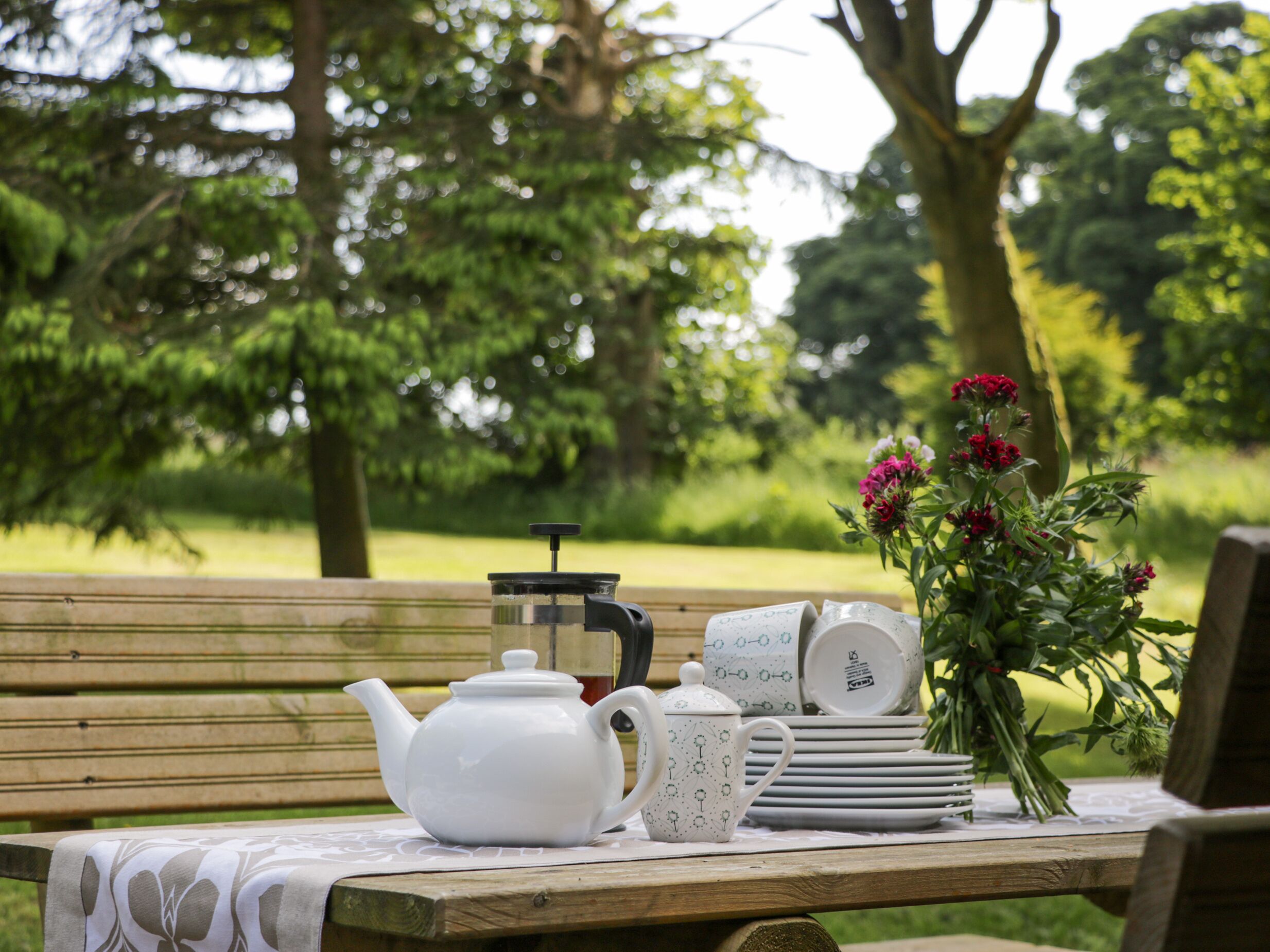 A picnic setup with a teapot, cups, and a vase of flowers on a wooden table in a park.